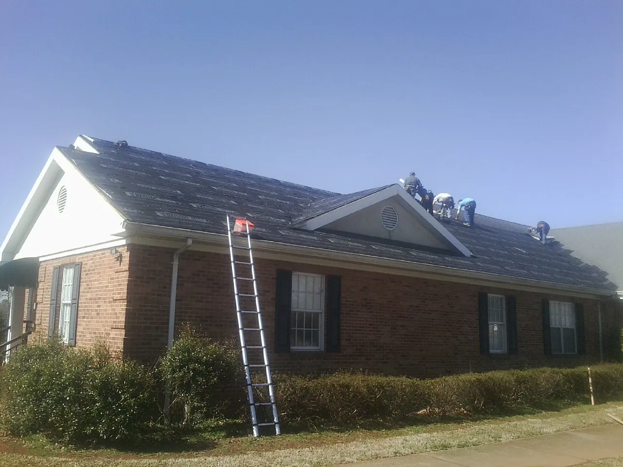 Crew tearing shingles off a brick ranch roof with a ladder set — Emergency Roof Repair work by our crew in Van Etten
