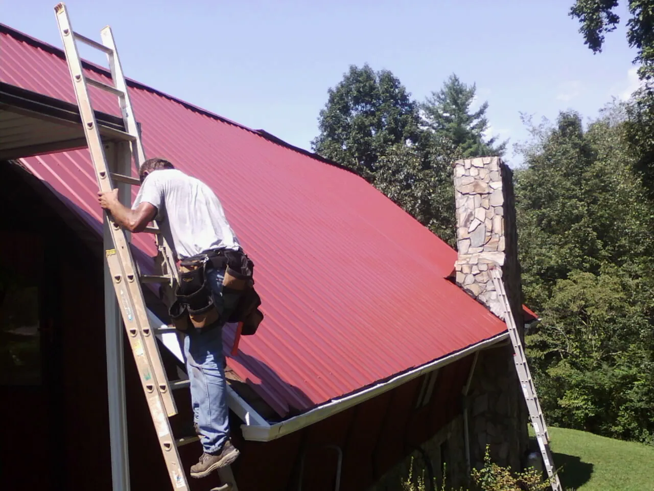 Roofer on a ladder beside a red metal roof and stone chimney — Soffit & Fascia Repair by Roofing Brothers in Van Etten